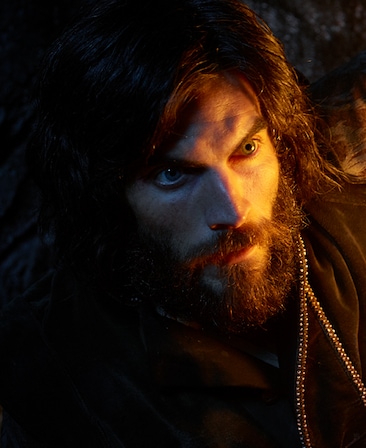 Wes Bentley headshot with beard and long hair in black top decorated with bejeweled detailing ends and half face illuminated