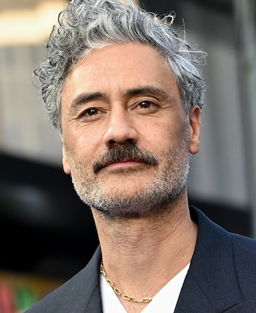 Taika Waititi headshot wearing white shirt with a black suit jacket and gold chain necklace.