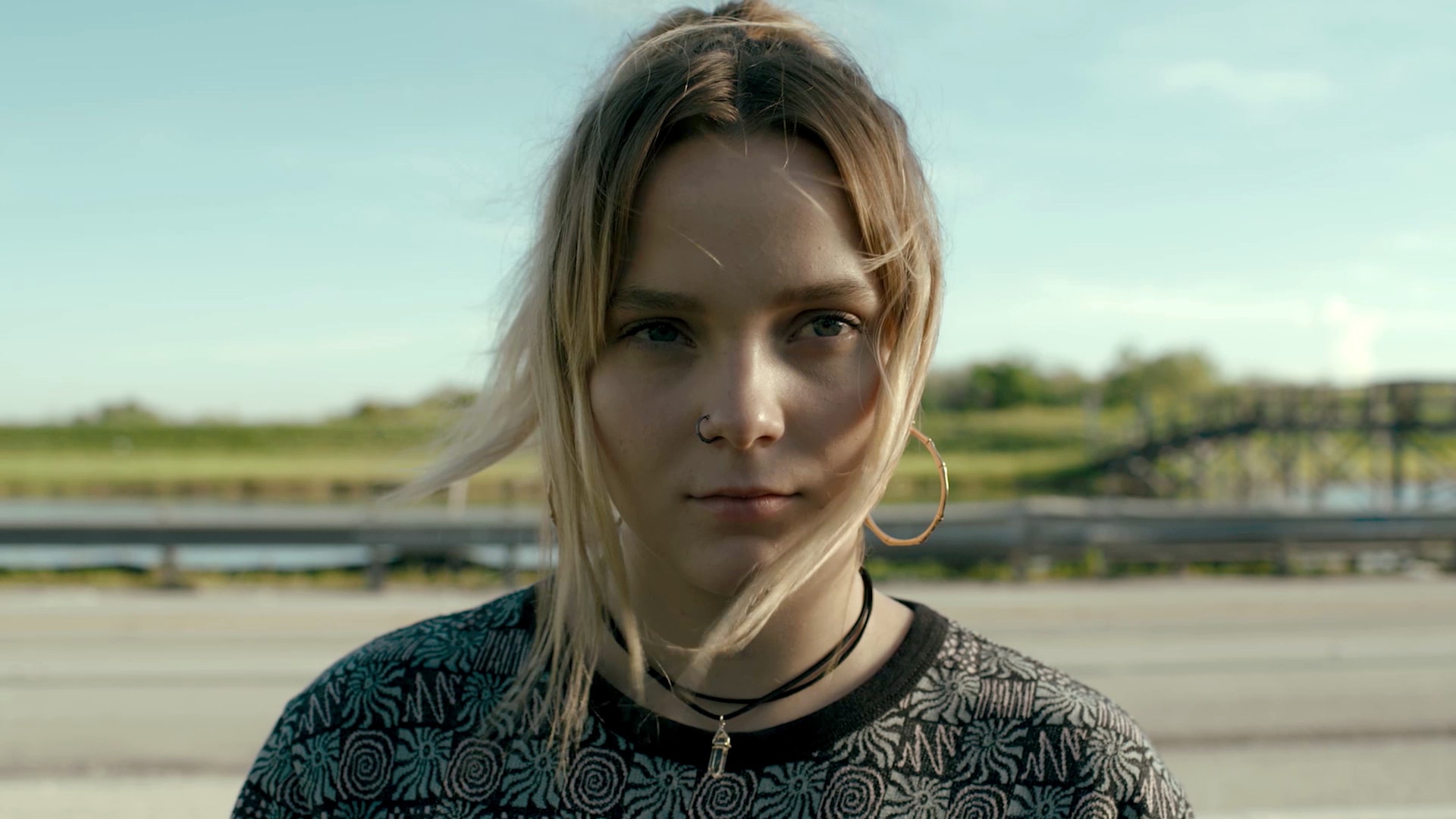 Willa Givens wearing a black choker necklace and patterned shirt, standing outside by a highway road