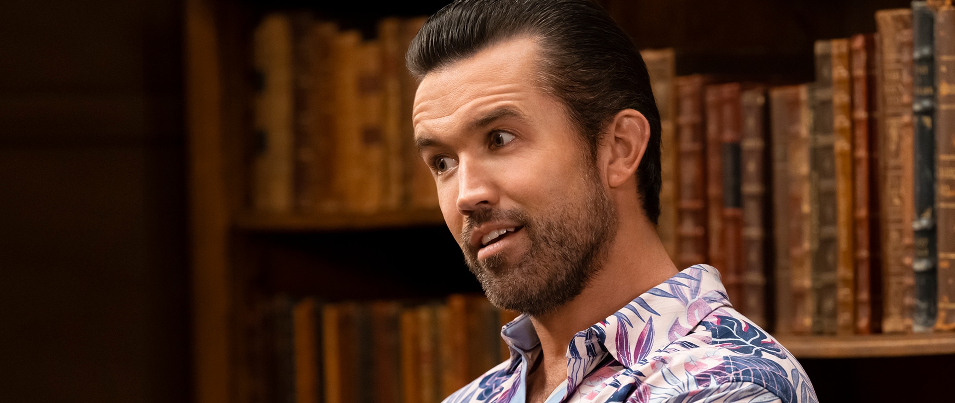 Rob McElhenney as Mac in FX's It's Always Sunny in Philadelphia wearing a tropical shirt with a bookcase behind him.