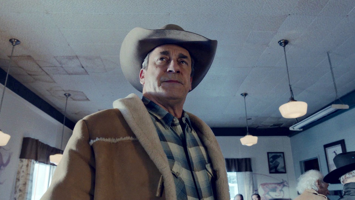 Man wearing a cowboy hat and blue flannel shirt with a brown coat, standing in a diner