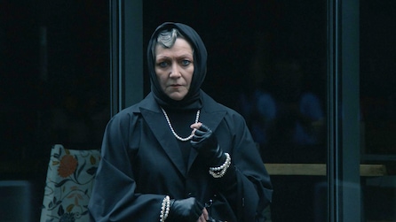 Woman in a black coat with a scarf wrapped around her head and wearing pearls