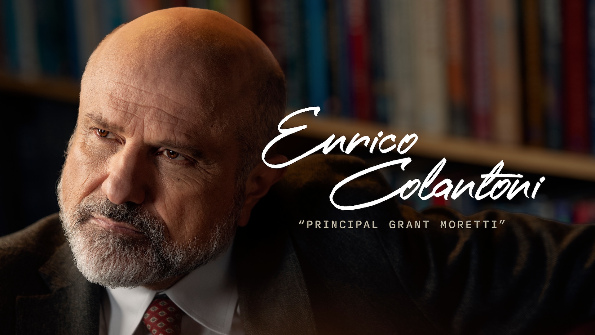 Enrico Colantoni as Principal Grant Moretti in FX's English Teacher, in a brown suit and red tie, and a bookshelf behind him.