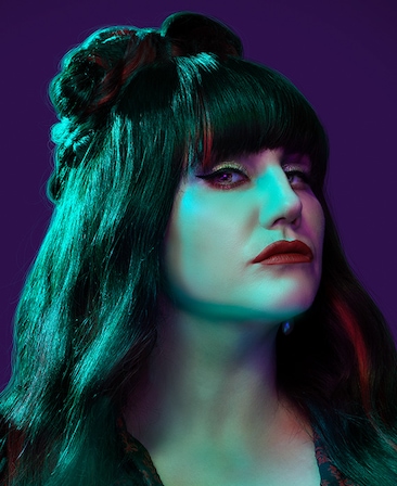 Natasia Demetriou headshot wearing red lipstick and standing in front of a purple background.