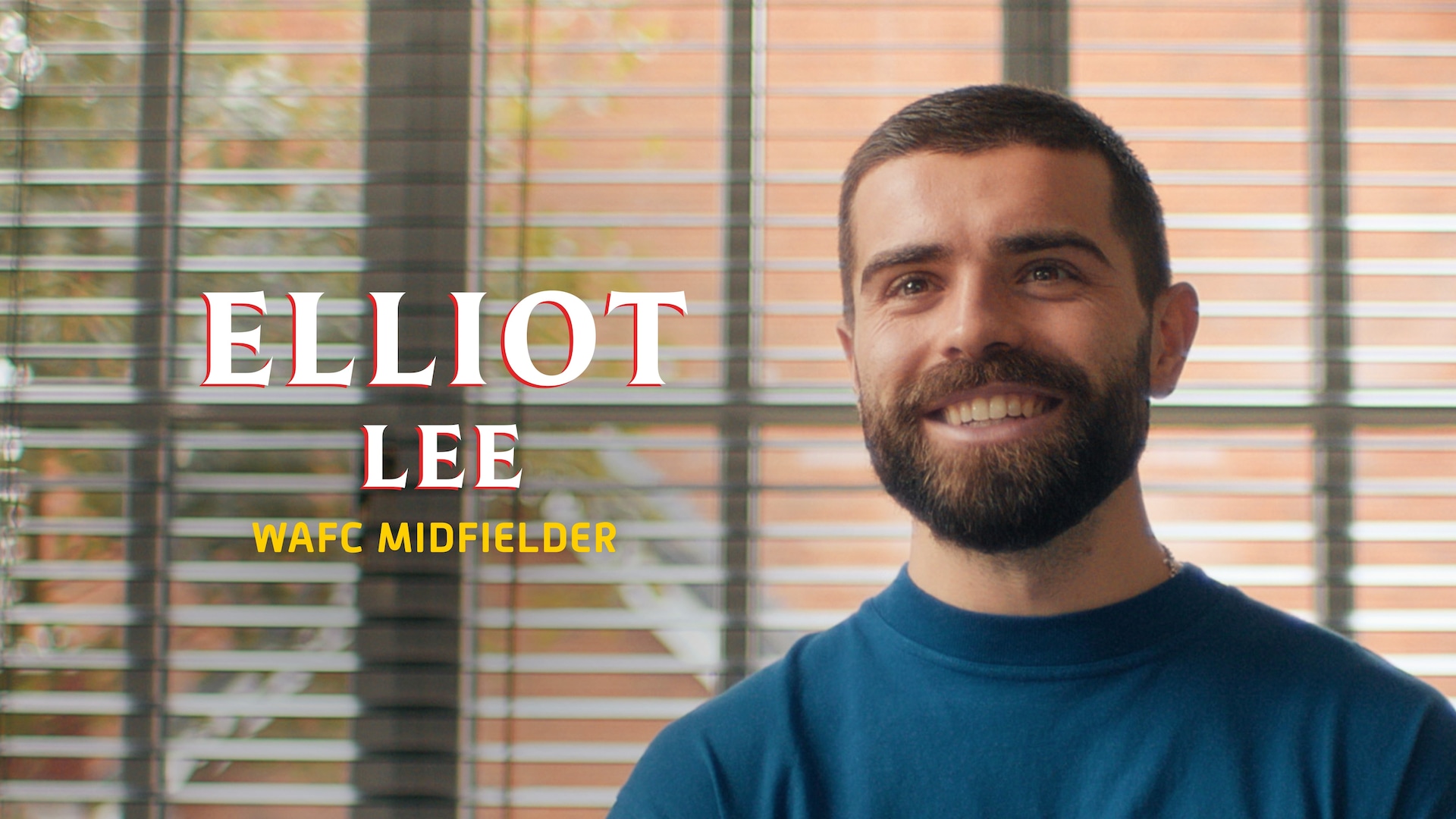 Elliot Lee headshot wearing a blue t-shirt, standing in front of a window