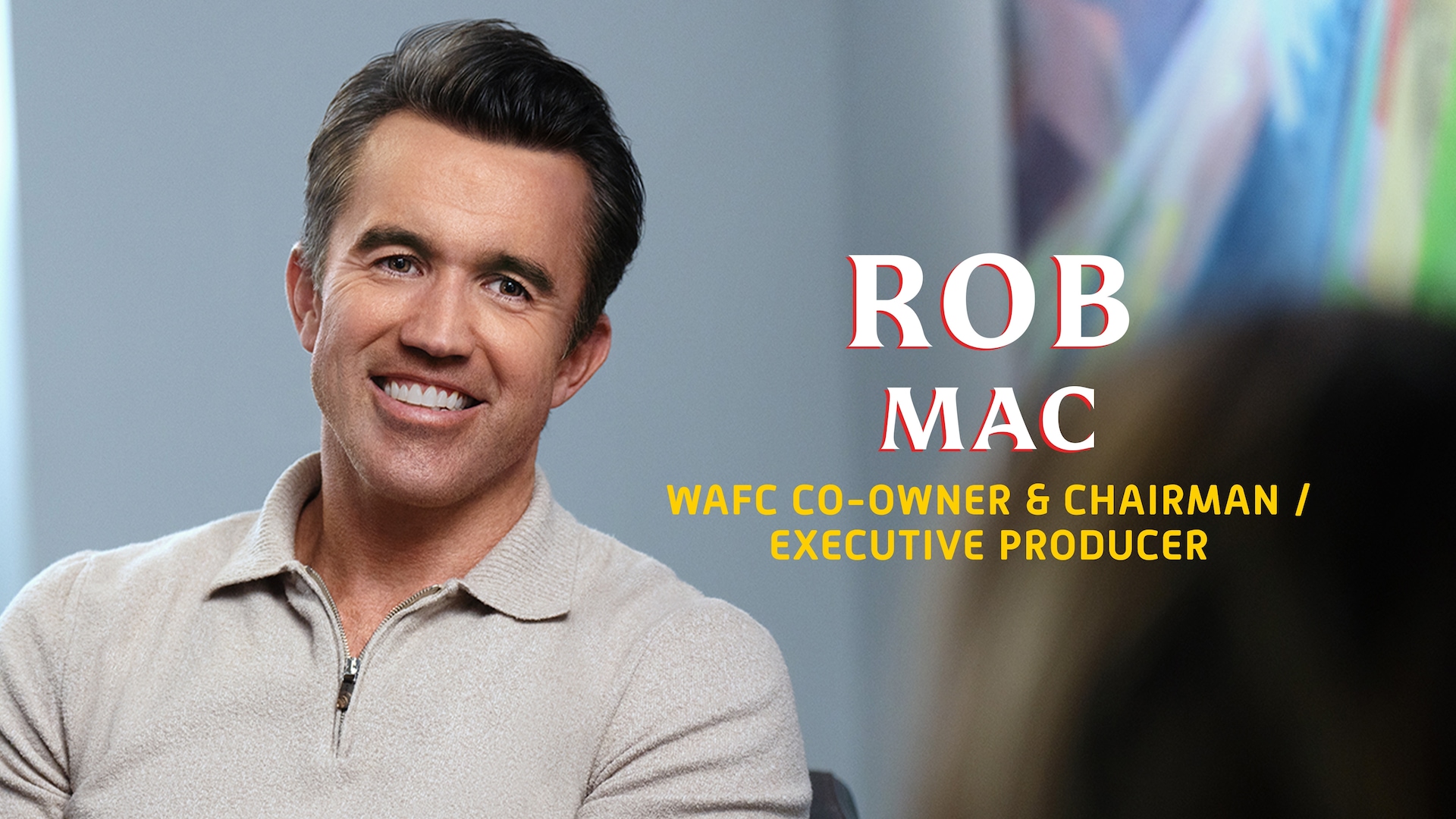 Rob Mac headshot wearing a beige zip up sweater whit his name in large white and red text to the side of him.