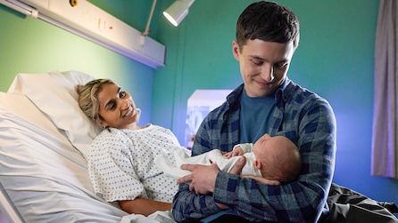 Luke holding a baby and Maya wearing a hospital gown and laying in a hospital bed.