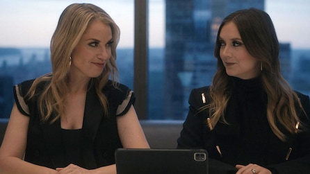 Two women wearing black tops sitting next to each other with a large window in the background and a view of buildings