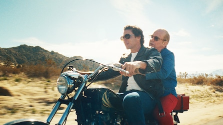 Truman Capote and a man with a leather jacket riding on the back of a motorcycle in the desert.