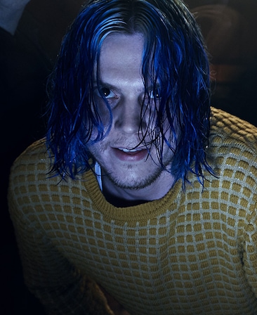 Evan Peters with wet long blue dyed hair staring up behind hair and leaning over in yellow and white square patterned sweater