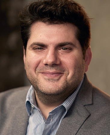Dan Shotz headshot wearing a blue checkered button up with a grey suit