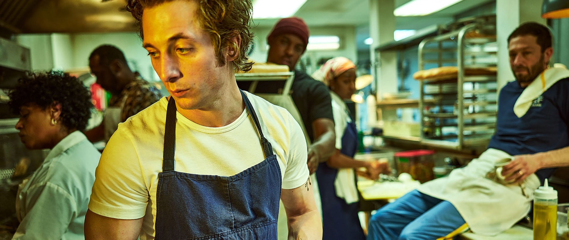 Carmy wearing a white shirt and blue apron in the kitchen of his restaurant.