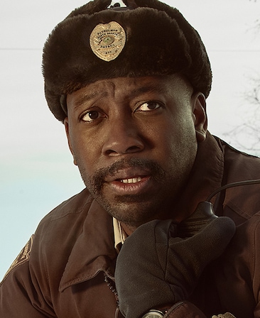 Lamorne Morris headshot wearing a fur hat with a sheriff badge on the front and a brown jacket with gloves