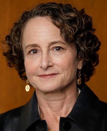 Nina Jacobson wearing a black satin button up wearing gold jewelry with a brown background