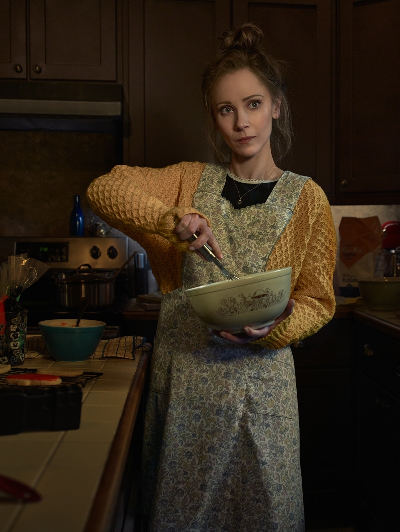 Woman with yellow sweater and blue floral apron standing in a kitchen while whisking in a bowl.