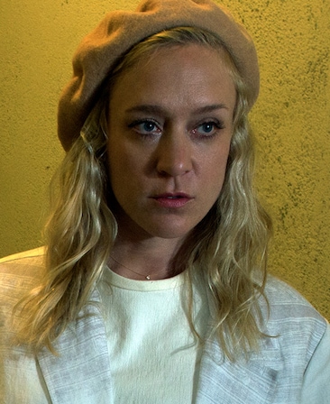 Chloe Sevigny headshot in brown beret against yellow textured wall, wearing white top and white coat