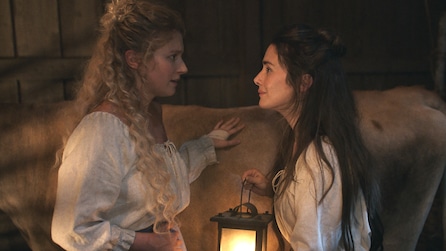 Two women in flowy white tops holding a lantern and petting a cow talking together in American Horror Stories Installment 2