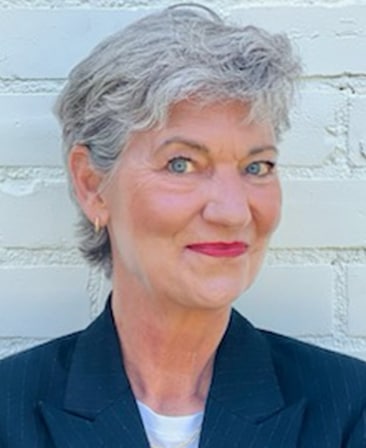 Headshot of Zanne Devine wearing a blue suit in front of a white brick wall.