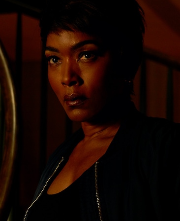 Angela Bassett headshot in black top and black zippered jacket in dark room with stairs in background and face illuminated
