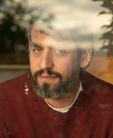Zal Batmanglij headshot wearing a red sweater sitting behind a window