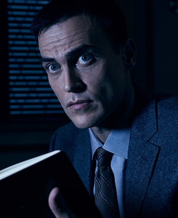 Cheyenne Jackson in grey suit and striped tie, looking up and holding black notebook in dark room with face illuminated