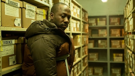 Man wearing brown and green jacket leaning on crutches in front of evidence boxes on shelves.