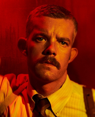 Headshot of Russel Tovey with mustache and striped shirt with suspenders under red light