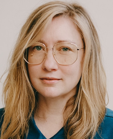 Headshot of executive producer Elizabeth Meriwether, wearing a blue shirt and wire framed glasses. 