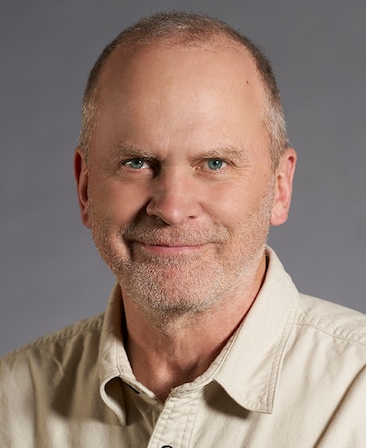Sam Johnson headshot wearing a beige button down shirt, standing in front of a gray background.