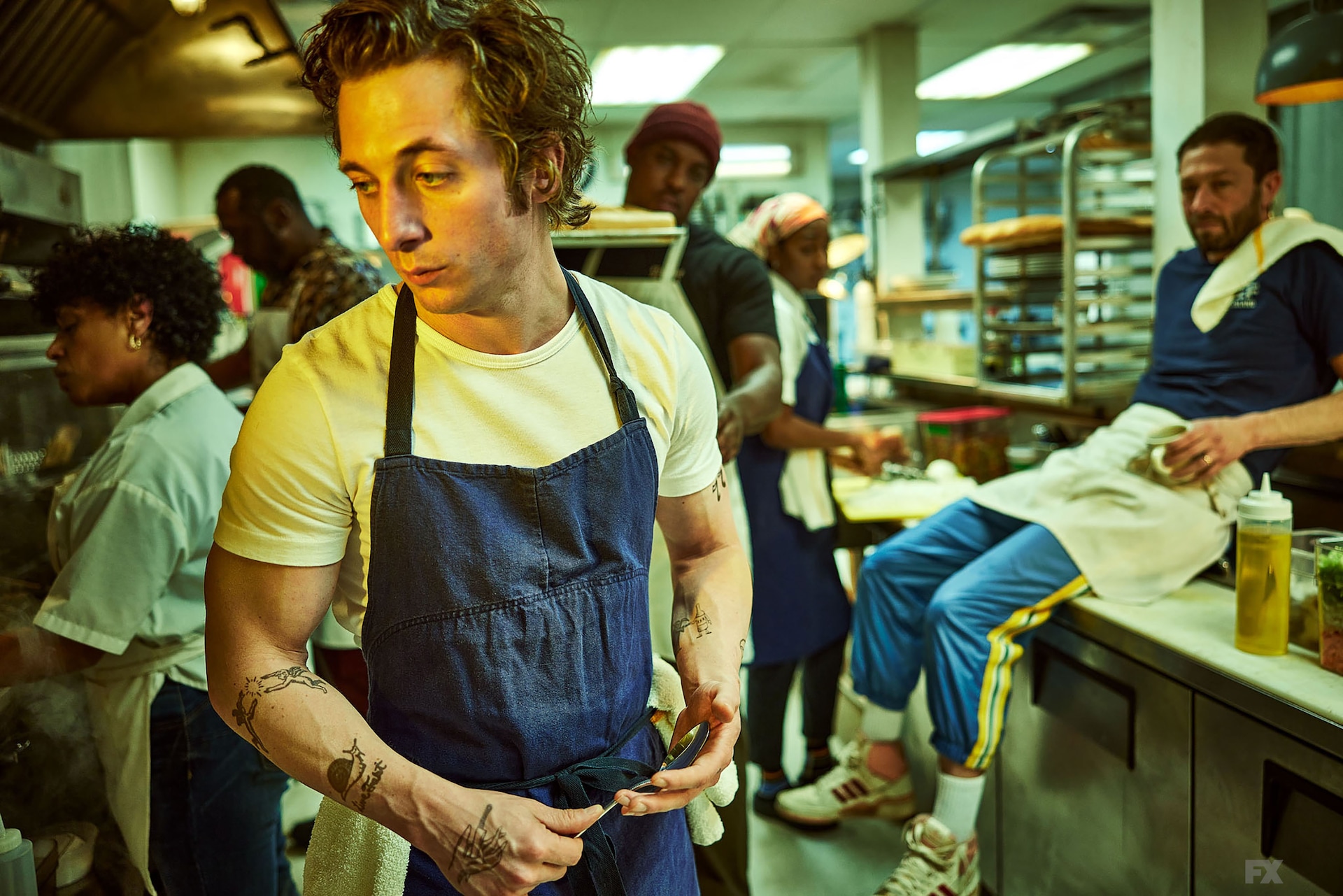 Jeremy Allen White as Carmy wearing a white shirt and blue apron while standing in the kitchen