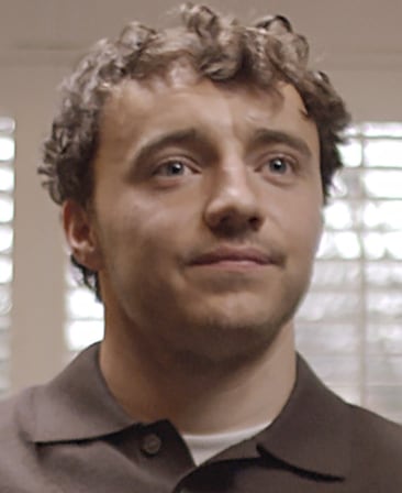 Ollie Rathbone headshot, wearing a brown button up with a white shirt peaking underneath, standing in front of window blinds.