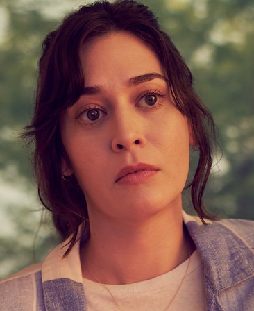 Lizzy Caplan headshot in blue and white checkered top with white shirt beneath, and blurred tree leaves in background