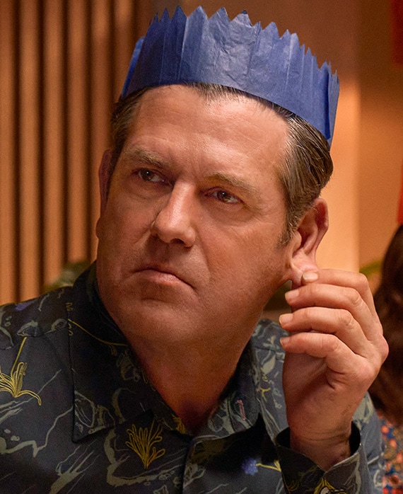 Patrick Baladi headshot wearing a blue paper crown and tugging at his ear with his fingers.