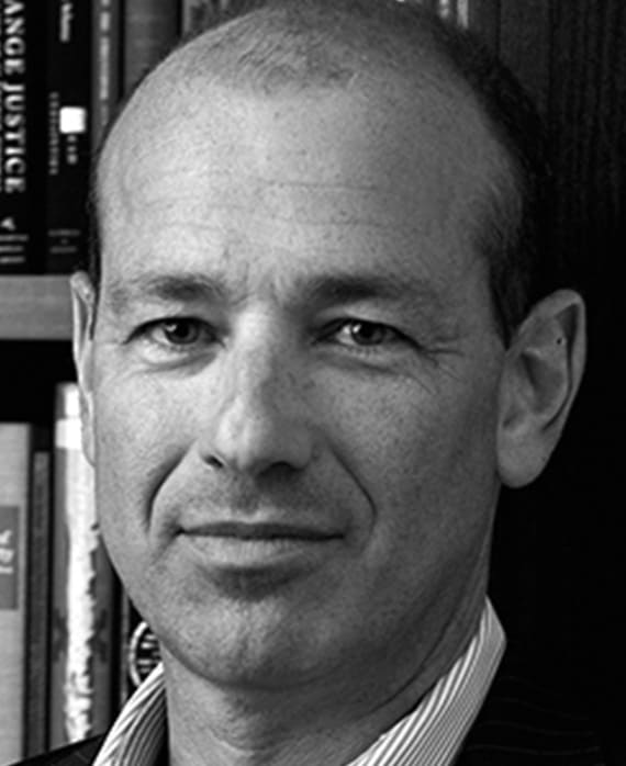 Howard Gordon headshot wearing a striped collar shirt in front of a bookshelf