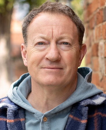 Simon Beaufoy headshot wearing a gray hoodie and plaid jacket, standing by a brick wall outside