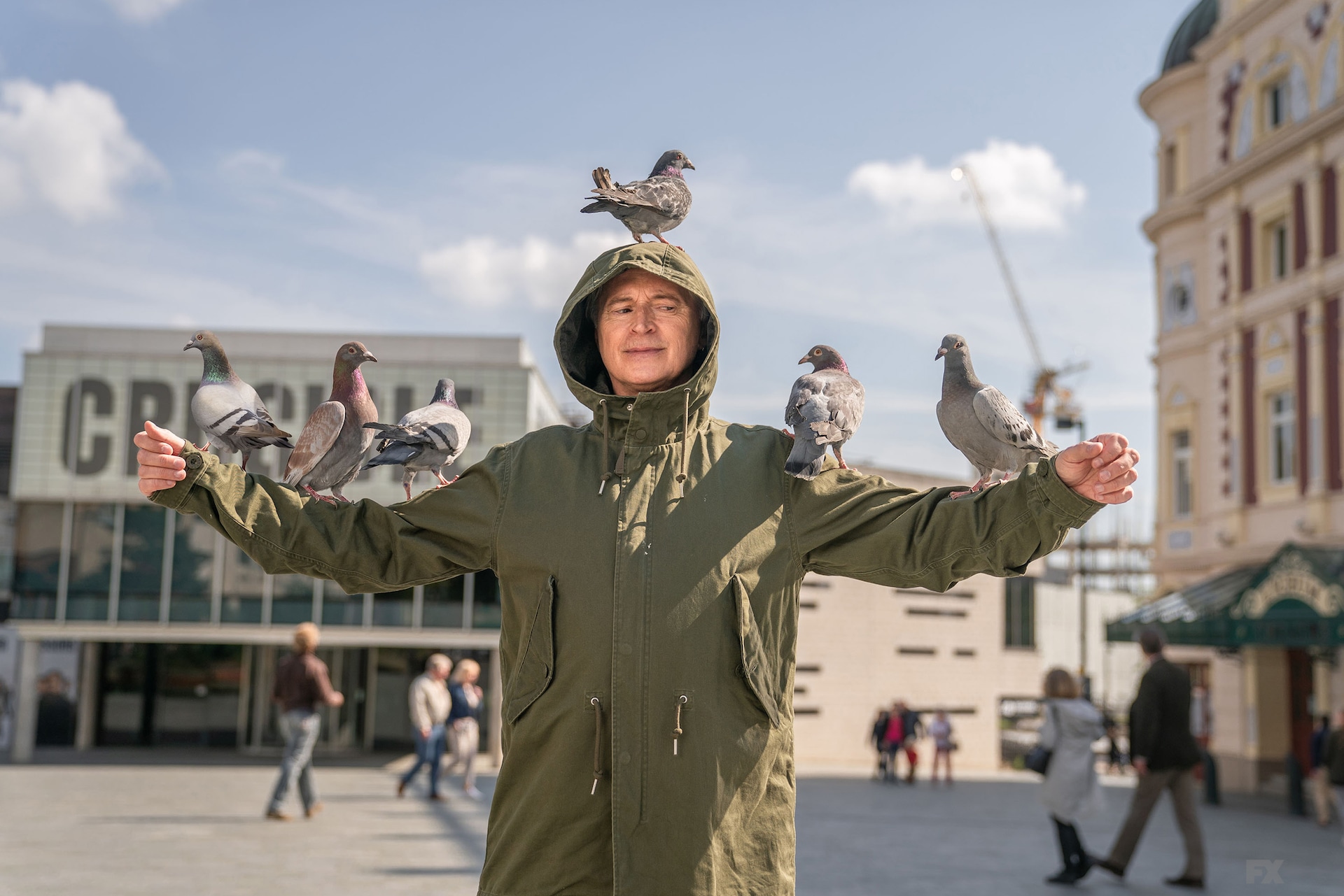Gaz wearing a green jacket with a hood and pigeons sitting on his arms and head