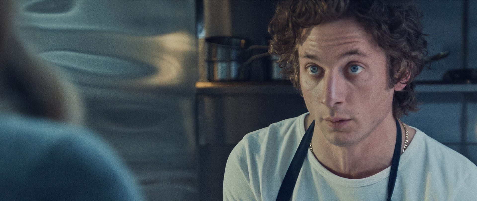 Carmy wearing a white shirt and blue apron in the kitchen of his restaurant