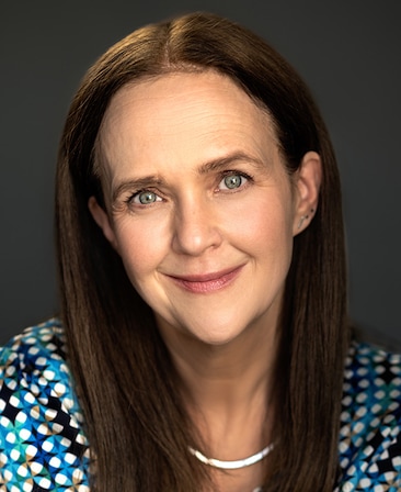 Headshot of Yana Gorskaya wearing a blue and black printed shirt and silver jewelry, in front of a grey background.