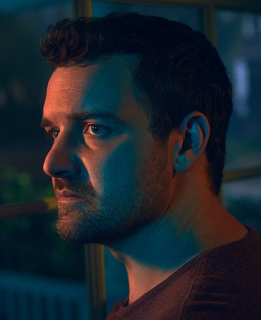 Micah Stock headshot of side profile, standing in front of house window at night with soft peach light shining on top of face