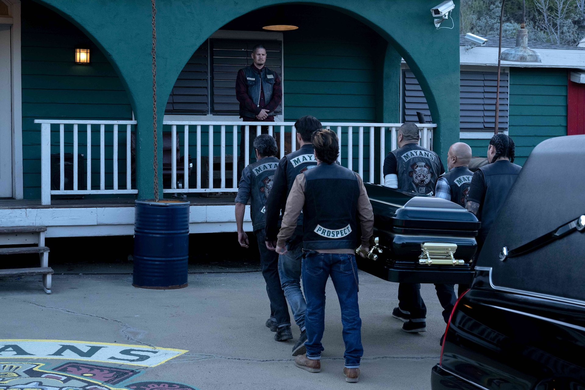 Mayans M.C. members carry a coffin from a hearse as EZ Reyes watches over them from a porch