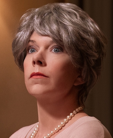 Mary Elizabeth Ellis headshot wearing a pink shirt, a pearl necklace, and a short silver wig.