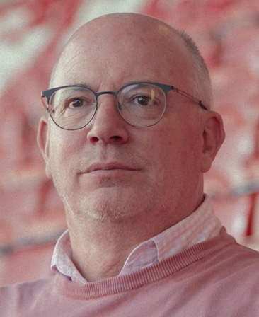 Shaun Harvey headshot wearing a pink sweater with a pink checkered button up underneath, sitting in the stands.