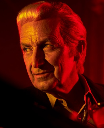 Headshot of Denis O'Hare as Henry in black suit and red lighting on face