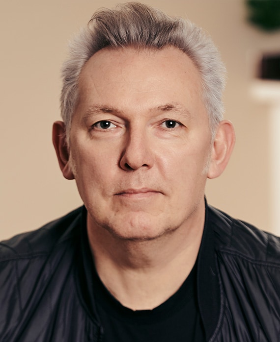 Richard Allen-Turner headshot in black shirt and black bomber jacket with peach colored background