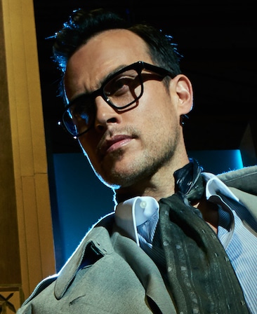 Cheyenne Jackson headshot in grey jacket with blue striped button down and dark grey scarf and glasses