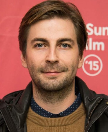 Jon Watts headshot wearing a brown sweater and black leather jacket standing in front of a red sign
