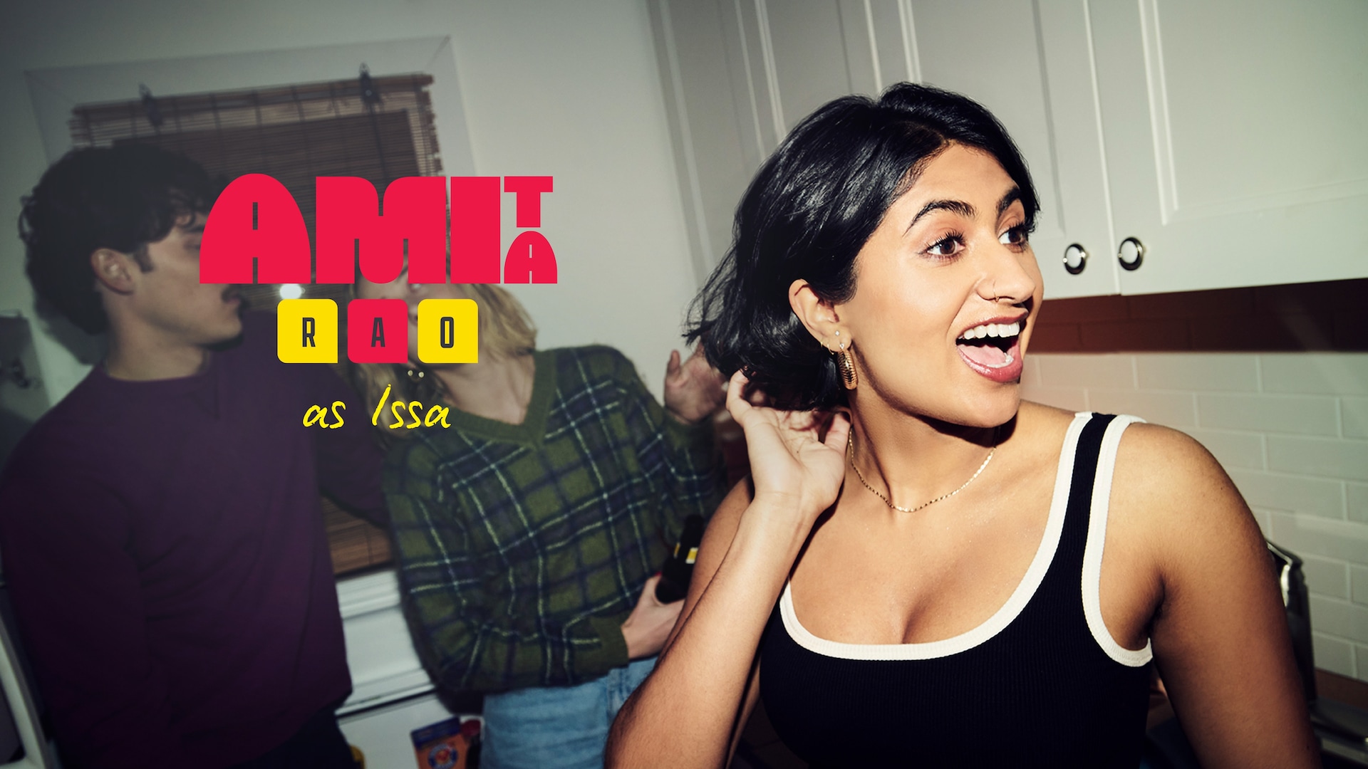 Amita Rao as Issa in FX's Adults, wearing a back tank top lined in white, smiling in a kitchen.