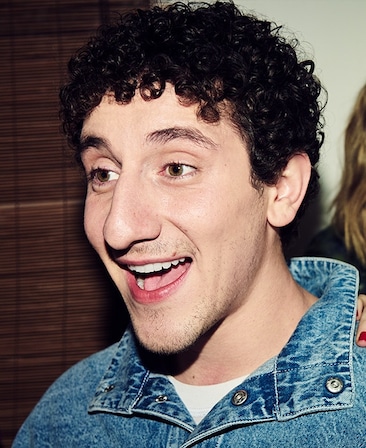 Headshot of Malik Elassal as Samir in FX's Adults, wearing a denim jacket and white shirt smiling for a flash photo.