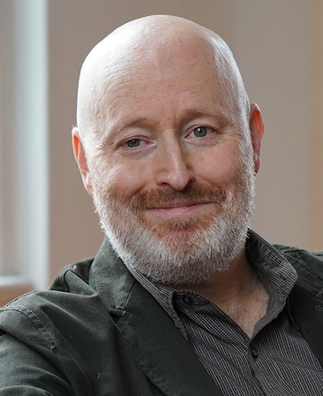 Joe Weisberg headshot wearing a button up shirt and green jacket
