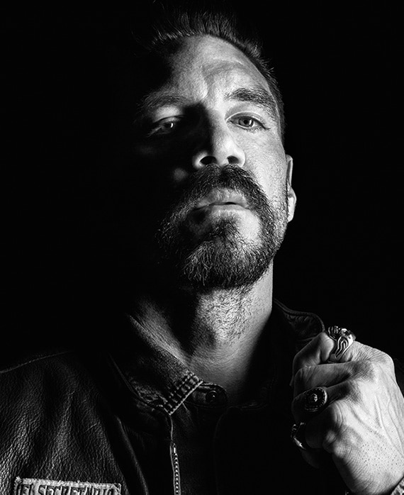 Clayton Cardenas headshot making a fist with his hand, wearing rings on his finger and a denim shirt